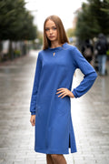 Long blue knit dress with flared silhouette, soft cotton fabric, decorative stitched flower detail on the side, elegant standing collar with a small front opening, loose sleeves gathered at the wrist, practical pockets, and one side slit. Mėlyna trikotažinė suknelė su skeltuku, kišenėmis ir dekoruota siuvinėta gėle.