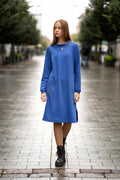 Long blue knit dress with flared silhouette, soft cotton fabric, decorative stitched flower detail on the side, elegant standing collar with a small front opening, loose sleeves gathered at the wrist, practical pockets, and one side slit. Mėlyna trikotažinė suknelė su skeltuku, kišenėmis ir dekoruota siuvinėta gėle.