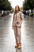 Woman wearing a cappuccino-colored two-piece leisure set with a hood and zipper. The sweatshirt has an asymmetric cut, front pockets, and a hand-decorated face motif. The trousers are straight and comfortable with an elastic waistband and pockets. Modern, cozy, and stylish outfit for everyday wear. / Moteriškas kapučino spalvos kostiumas su gobtuvu, dekoru ir kišenėmis – stilingas ir patogus kasdienai.