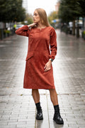 Woman wearing a terracotta velvet (corduroy) dress with long sleeves gathered at the cuffs, side pockets, and a subtle neckline detail. The dress is made from a soft cotton-based fabric with fine stripes. Mid-length, breathable and elegant design for autumn and winter. / Terakotos spalvos veliūrinė suknelė su kišenėmis ir subtiliu kaklo dizainu, iš medvilninio audinio su juostelėmis.