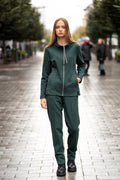 Comfortable dark green women's two-piece tracksuit made of soft brushed-inside cotton jersey; features a full-zip hoodie with decorative stitched pockets, high collar, subtle side slits and straight-cut trousers with an elastic waistband. The set is designed for everyday wear, offering warmth, softness and a clean silhouette. / Tamsiai žalios spalvos moteriškas trikotažinis laisvalaikio kostiumėlis su švelniu pūkeliu viduje, dekoratyvinėmis kišenėmis, aukšta apykakle, užtrauktuku ir tiesaus kirpimo kelnėmis