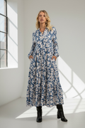 Ilga mėlyna gėlėta moteriška maxi suknelė iš 100 % viskozės su ilgomis rankovėmis, sagomis priekyje ir subtilia stovinčia apykakle, lengvai platėjančio silueto su raukta apačia, dekoruota smulkiais baltais gėlių žiedeliais mėlyname fone, elegantiška ir patogi kasdienai bei laisvalaikiui.
