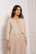 Smėlinės spalvos bolero tipo kardiganas su subtiliomis lurex juostelėmis, 3/4 rankovėmis ir viena užsegimo sagute, dėvimas ant lengvos puošnios suknelės, elegantiškas ir švelnus moteriškas įvaizdis šviesiame interjere. / Beige sand bolero cardigan with subtle lurex stripes, 3/4 sleeves and single button fastening, styled over a delicate dress, elegant feminine look in a bright interior.