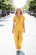 Woman wearing a bright yellow two-piece linen set on a city street. The outfit features a short sleeve top with handcrafted front detail and wide-leg trousers.