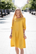 Bright yellow linen dress with floral embroidery, bell-shaped silhouette, side pockets and folded sleeves. Model wearing the dress outdoors in summer sunlight.