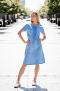 Light blue linen summer dress for women made from 100% softened linen, featuring a relaxed silhouette, short sleeves, two side pockets, and a delicate handmade-like decorative front detail. Ideal for warm weather, breathable and stylish. Modelis vilki melsvą lininę suknelę trumpomis rankovėmis, tinkamą vasarai – natūralus audinys, patogumas ir elegancija viename.