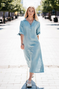 Lininė suknelė prigesintos mėtos spalvos su kišenėmis ir dekoratyvinėmis gėlėmis – moteris vaikšto gatve. / Muted mint linen dress with floral embroidery and pockets – woman walking in the street.