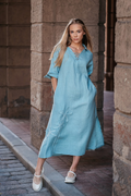 Lininė suknelė prigesintos mėtos spalvos su kišenėmis ir dekoratyvinėmis gėlėmis – moteris vaikšto gatve. / Muted mint linen dress with floral embroidery and pockets – woman walking in the street.