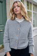 Pilkos spalvos angoros mišinio moteriškas kardiganas su ilgomis rankovėmis, apvalia kaklo iškirpte, priekyje esančiomis kišenėmis ir dekoratyvinėmis auksinėmis sagomis, minkštas ir šiltas mezginys, lengvai derinamas prie kasdienio ir elegantiško stiliaus, tinkamas rudens ir žiemos sezonui