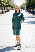 Woman wearing a dark green linen dress with 3/4 sleeves and school-style collar, standing on a sunny street. / Moteris dėvi tamsiai žalią lininę suknelę su 3/4 rankovėmis ir mokykline apykakle, stovi miesto gatvėje.

Close-up of front design showing decorative seams and soft linen fabric. / Artimas vaizdas į priekį – matomos išryškintos siūlės ir minkštas lino audinys.

Side view of the linen dress with subtle silhouette and front slit. / Šoninis vaizdas – subtilus siluetas ir skeltukas priekyje.